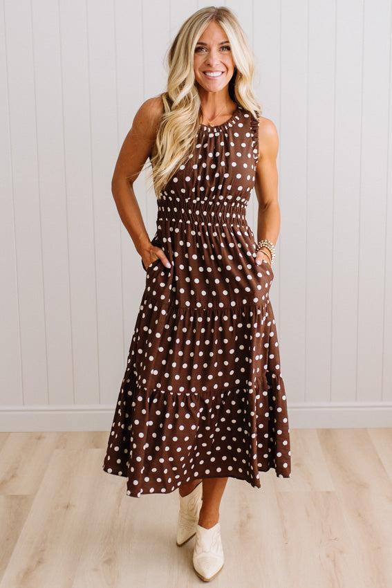 A woman modeling a sleeveless brown midi dress with white polka dots, featuring a smocked bodice and side pockets, paired with cream ankle boots against a white paneled wall and light wood floor.