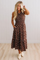A woman modeling a brown sleeveless midi dress with white polka dots, featuring a smocked waist and side pockets, paired with cream ankle boots against a white paneled wall and light wood floor.