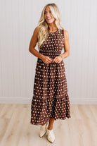 A woman modeling a sleeveless brown midi dress with white polka dots, paired with white pointed-toe boots, standing in front of a white paneled wall and light wood floor.