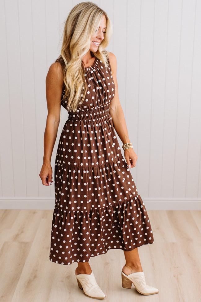 A woman modeling a brown sleeveless midi dress with white polka dots, paired with cream-colored clogs, standing in front of a white paneled wall and light wood floor.