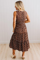 A woman wearing a sleeveless brown midi dress with white polka dots, shown from the back against a white paneled wall and light wood floor.
