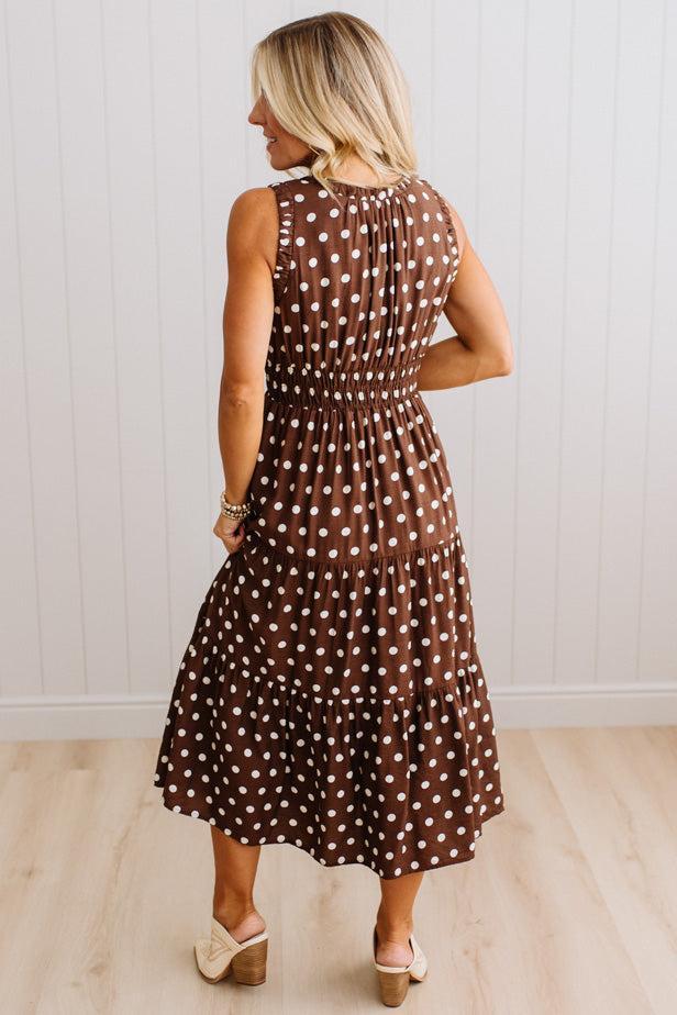A woman wearing a sleeveless brown midi dress with white polka dots, shown from the back against a white paneled wall and light wood floor.