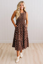 A woman modeling a sleeveless brown midi dress with white polka dots, featuring a smocked bodice and side pockets, paired with white ankle boots against a white paneled wall and light wood floor.