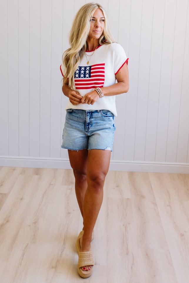 Woman wearing a white t-shirt with an American flag design and denim shorts indoors.