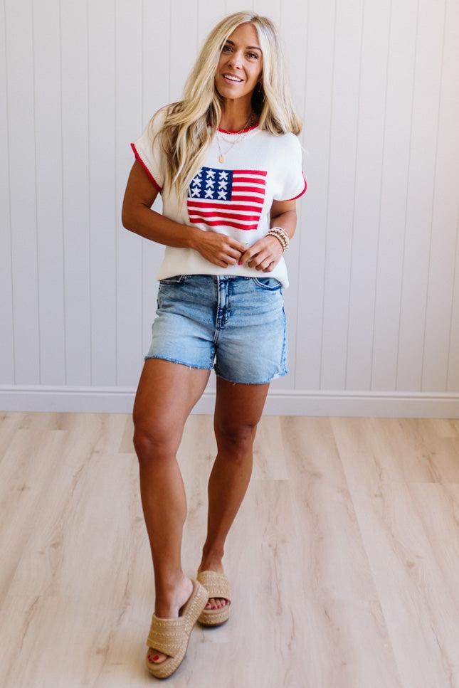 Woman wearing a white t-shirt with an American flag design and denim shorts indoors.