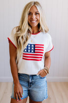 Woman wearing a white t-shirt with an American flag design and denim shorts indoors.