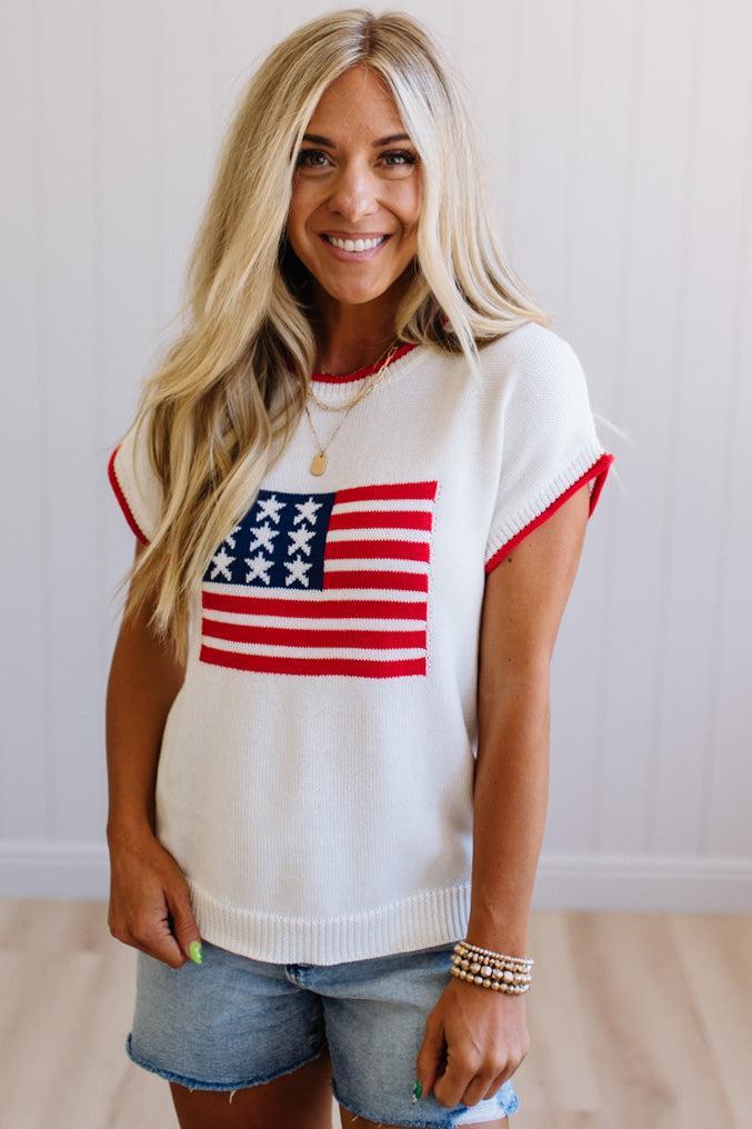 Woman wearing a white sweater with an American flag design, standing against a plain background.