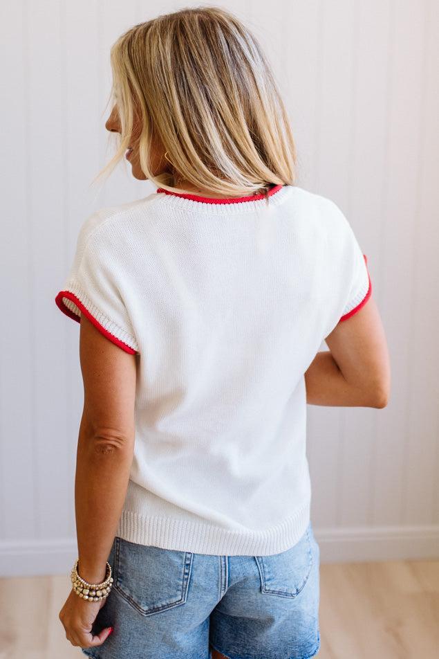 Woman wearing a white sweater with red trim and blue jeans against a plain background
