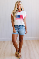 Woman wearing a white t-shirt with an American flag design and denim shorts indoors.