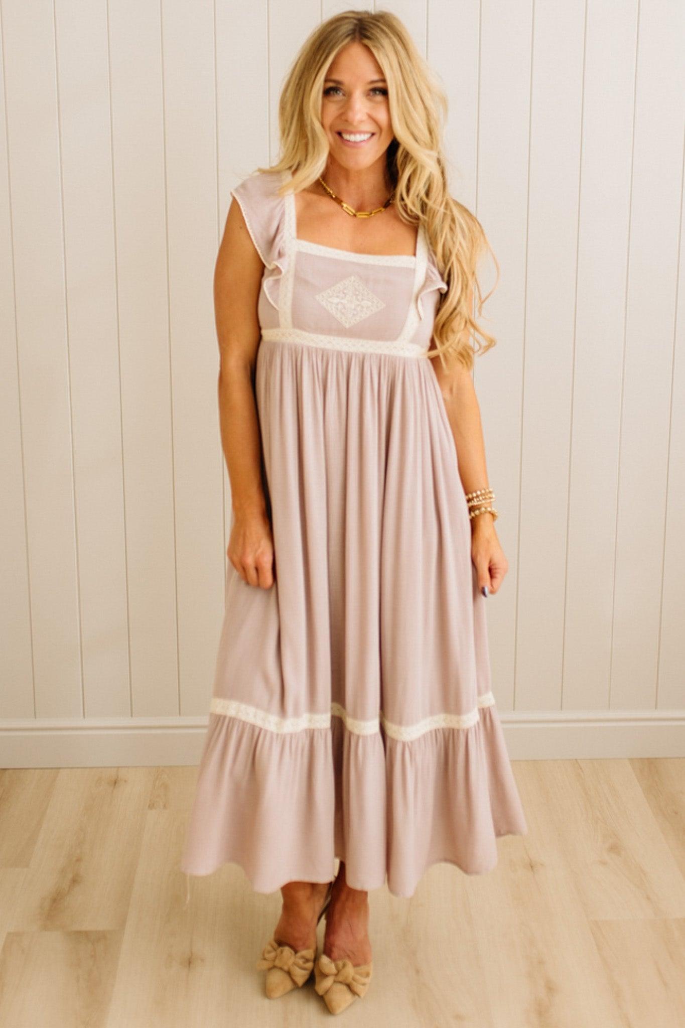 Woman wearing a long, light-colored dress with a white top against a wooden paneled wall.