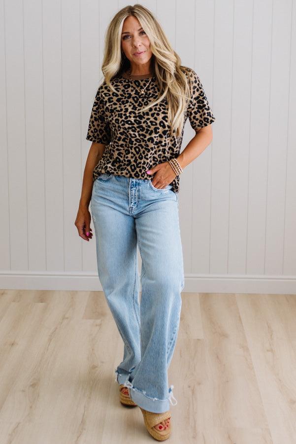 Woman wearing a leopard print top and light blue jeans standing against a white paneled wall.