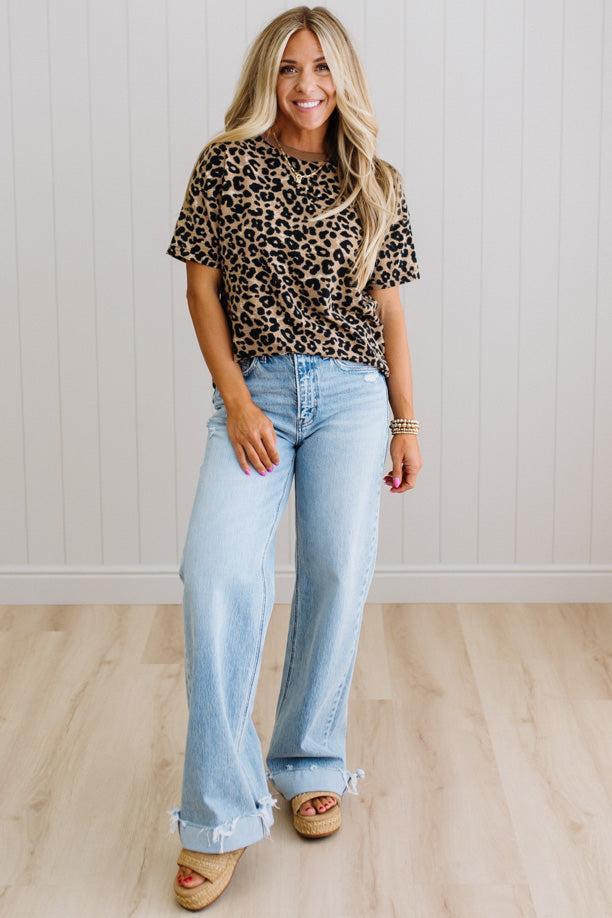 Woman wearing a leopard print top and light blue jeans standing against a white paneled wall.