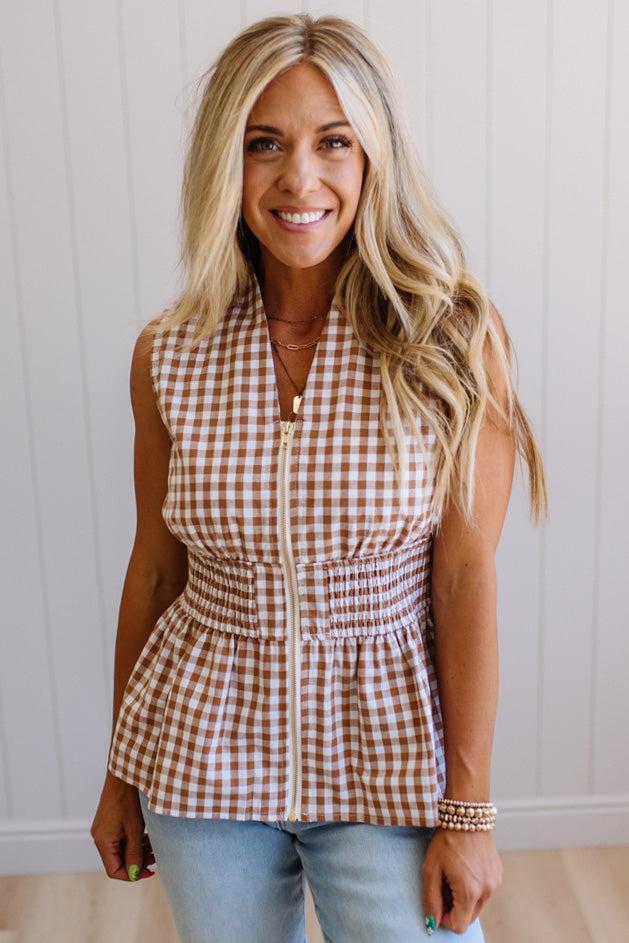 Blonde woman wearing a mocha and white gingham print sleeveless top with smocked waistline and centered gold zipper front.