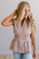 Blonde woman wearing a mocha and white gingham print sleeveless top with smocked waistline and centered gold zipper front.