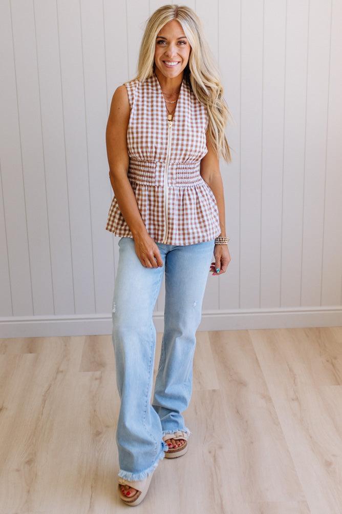 Blonde woman wearing a mocha and white gingham print sleeveless top with smocked waistline and centered gold zipper front.