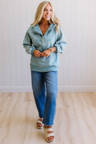 Woman wearing a light blue jacket and jeans standing against a white paneled wall.