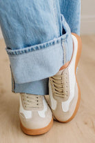 Beige sneakers with white accents and brown soles worn with light blue jeans on a wooden floor.
