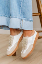 White and brown shoes worn with rolled-up blue jeans on a wooden floor.