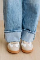 Close-up of rolled-up blue jeans and beige sneakers on a wooden floor.