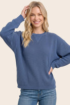 A woman wearing a blue ribbed knit crewneck sweater and blue jeans, posing against a solid light beige background.