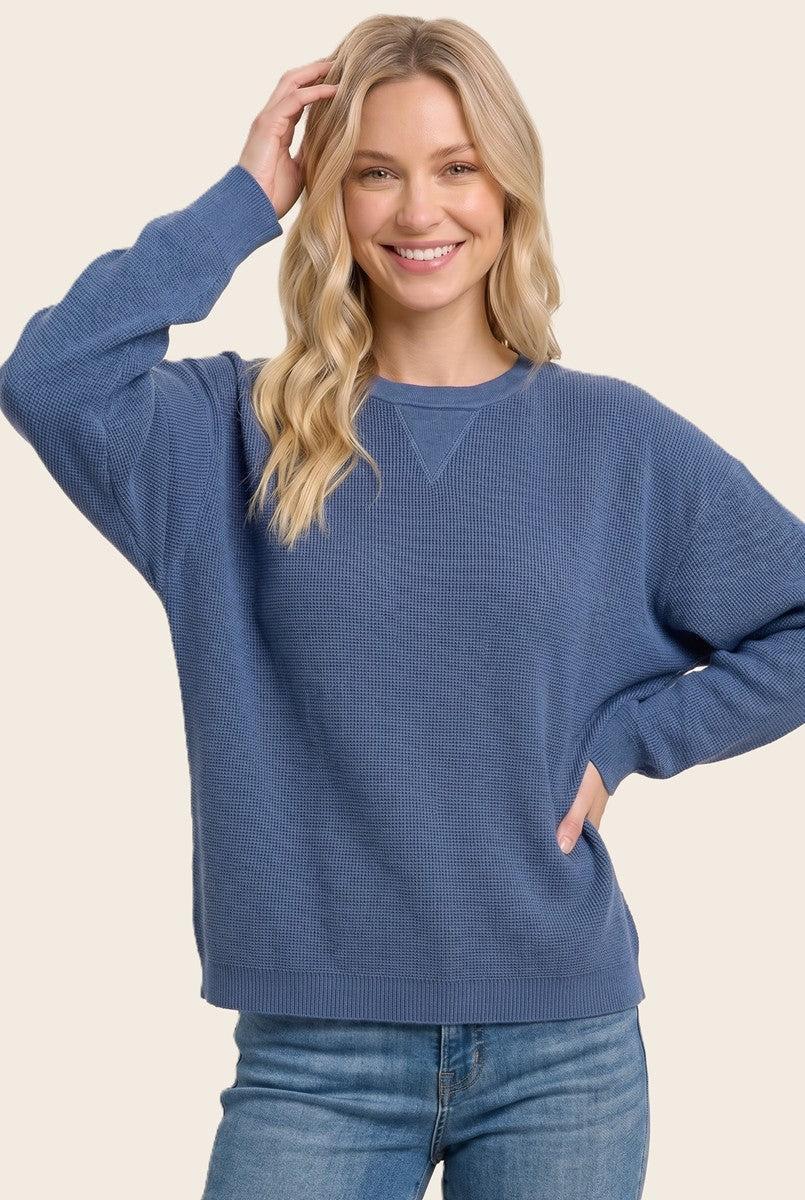 A woman wearing a blue ribbed knit crewneck sweater and blue jeans, posing against a solid light beige background.